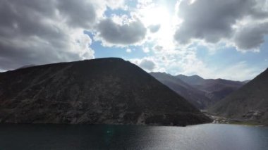 Derin Yeşil Göl 'ün sinematik hava perspektifi, Karanlık Dağlar tarafından çerçevelenmiş, bir yol kenarından görülen, Mavi ve Yumuşak Bulutlar ile dramatik bir gökyüzü altında,