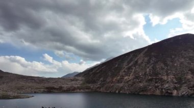 Görkemli Dağ Sıraları, Karanlık Dağlar, Mavi Gökyüzü, Beyaz Yumuşak Bulutlar, Kara Bulutlar, Hafif Yağmur Damlaları ve Huzurlu Karanlık Yeşil Suların keyfini çıkaran Turistlerle Görkemli Göl Manzarası.