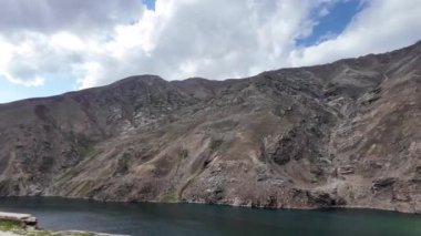 Sükunet Yeşil Göl 'e bakan virajlı bir yoldan yan görüntü. Çimenli ve Kayalıklı Karanlık Dağlar, Mavi Gökyüzü, kabarık bulutlar, Kara Bulutlar, Hareket Bulutları ve Hafifçe Düşen Işık Yağmurları