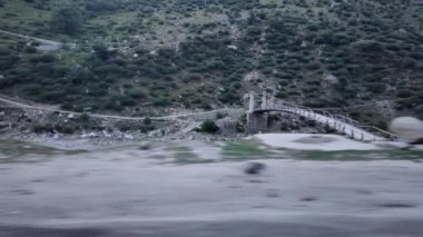 Bir yol kenarı manzarası, diğer tarafa giden yolu bağlayan bir köprüye sahip bir nehir içerir. Arka planda, görkemli bir dağ manzarası çimen ve ağaçlarla süslenir.