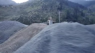 Hareket halindeki bir yol manzarasında beton yığını ve çakıl döküm alanı, büyük yeşil dağlar, elektrik kuleleri ve ağaçlardan oluşan bir zemin yer alıyor.