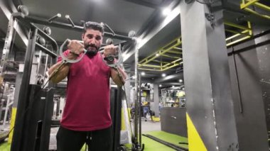 Spor salonunda Makine 'de Dururken İki Kolunda Kablo Kıvrımları Yapan Büyük Kas ve Görünür Damarlara sahip Vücut Geliştirici, Arka plan İnsanları Çalışıyor, Yan-Ön Görünüm