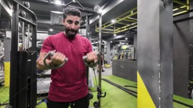 Vücut Geliştirici, Büyük Kaslı ve Görünür Damarlar, Spor Salonunda Makinede Dururken İki Kolunda da Kablo Biceps Kıvrımları Yapıyor, Saat Takıyor, Bileklik Takıyor, Bir Ön Kollu Dövme, Arka planda Çalışan İnsanlar,