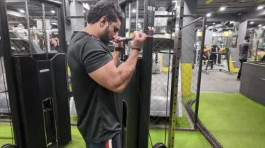 Bir Makinenin Üzerinde Dururken Kablo Bicep Kıvrımları Yapan Adam, Arka planda Egzersiz Yapan İnsanlarla Büyük Pazılarını ve Kaslarını Gösteriyor