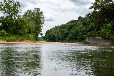 Parlak güneşli bir gün güzel, huzurlu bir nehir manzarası oluşturur. Sakin suların üzerinde yeşil ağaçlar ve beyaz bulutlar rahatlatıcı bir rahatlama hissi verir..