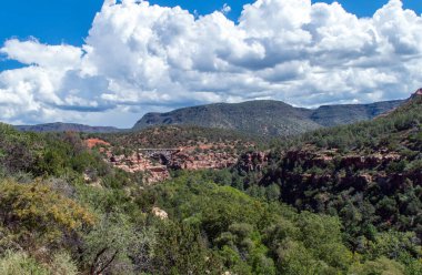 Yeşil çalı, kırmızı kaya, dağlar, mavi gökyüzü ve beyaz bulutlarla çevrili bir köprü Arizona 'dan güzel sakin bir manzara sunar..