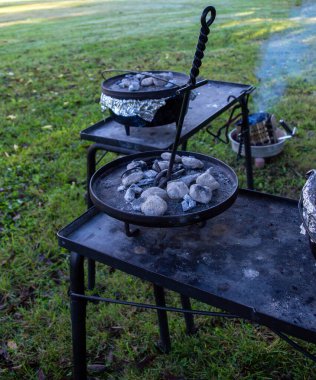 Açık havada dökme demir Hollanda fırını ve kömür briketleri kullanarak yemek pişirmek. Dumanlı kömürün kokusunu hayal edebilirsiniz ve tencerede ne tür lezzetli malzemeler olduğunu merak edebilirsiniz. Bokeh.