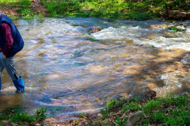 Plastik çizmeli tanımlanamayan bir adam arka plandaki kaya çıkıntısının üzerinden çağlayarak akan suları aşarak nazikçe akan bir derede yürüyor. Bokeh.