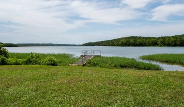 İnsan yapımı bir ayak köprüsü göl kenarı rıhtımının yolu. Önde yeşil çayır, ortada göl ve uzakta mavi gökyüzü ve bulutları olan ağaçlar..