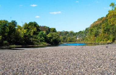 Kara çakıl taşları nehrin önündeki bir bölgeyi kaplar. Ağaçlar, su, mavi gökyüzü ve kayayla doğal bir sahne çeker. Bokeh.