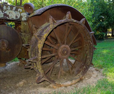 Arka planda hafifçe çözünmüş yeşil ağaçlarla yeşil çimlerde sergilenen ağır dökme demirden yapılmış yıpranmış eski bir traktör tekerleğine yakından bakın..