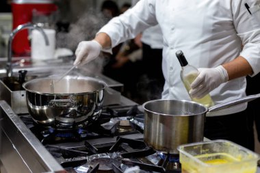 Mutfakta tencere karıştıran bir aşçı. Mutfakta büyük saksılar var.