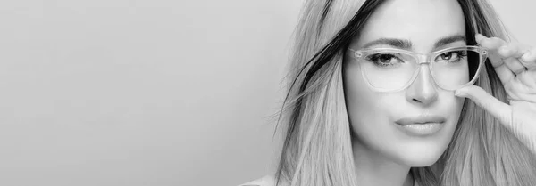 Monochrome fashion eyewear and clear vision concept. Close up studio portrait of a seductive blonde young woman wearing clear eyeglasses while looking at camera against grey background for copy space.