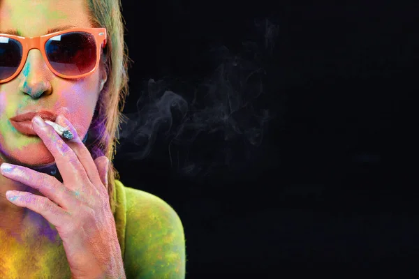 Beautiful woman covered in rainbow colored powder smoking weed. Close-up portrait isolated on a black panorama background with copyspace