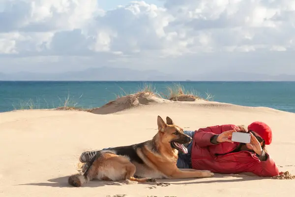 Photographer captures dog playing on beach with smartphone amidst scenic sand dunes.