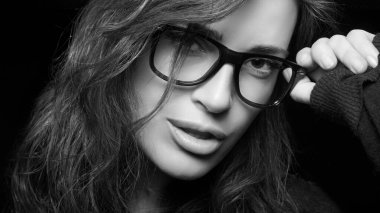 Close up of a young woman holding her eyeglasses while looking at camera. Cool trendy eyewear portrait isolated on black background