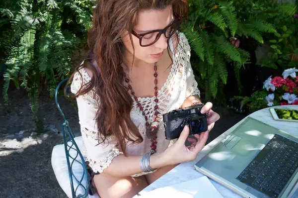 一个有着棕色长发 眼镜和自然发型的女人坐在户外 四周环绕着自然和开花的植物 她用笔记本电脑和照相机学习 把技术和闲暇结合起来 — 图库照片