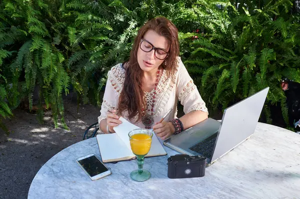 A woman in glasses works on a laptop in a park, enjoying the freedom and beauty of nature. She perfectly combines work and leisure.