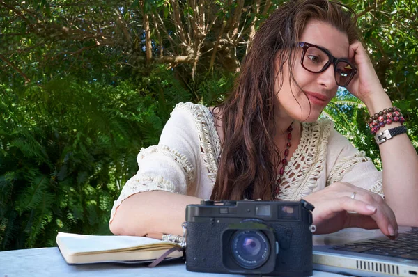 안경과 복고풍 스타일을 여자는 자연에 둘러싸인 노트북과 카메라와 야외에 앉아있다 — 스톡 사진