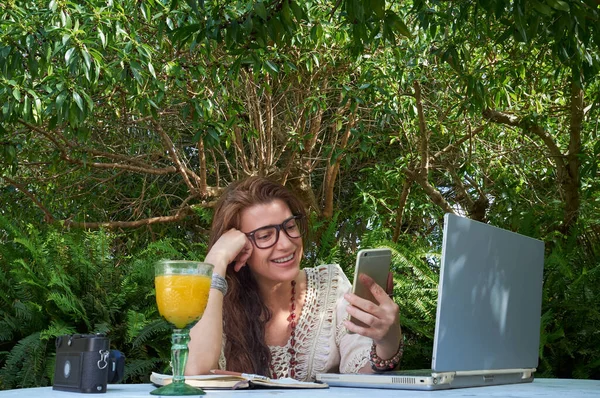 A cheerful woman works outdoors, enjoying a drink and using a vintage laptop, smartphone and camera. She sits at a table under a tree, smiling happily while balancing work and leisure.