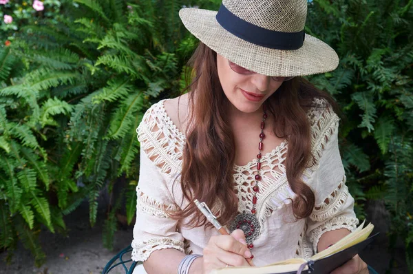 一个穿着休闲装的年轻姑娘在户外工作 她戴着太阳帽 在大自然的包围下学习 她的长发和草帽有助于她的游牧数字化生活方式 图库图片