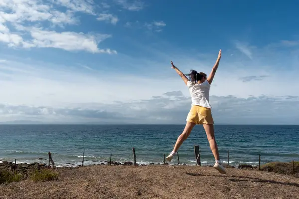 Confident woman jumps outdoors at the ocean shore, capturing a real moment of wellness and freedom.