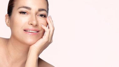 50 'lerin güzel doğal kadını gülümseyip yüzüne dokunuyor, cilt bakımını ve güzelliğini temsil ediyor. Pembe arkaplanda kopyalama alanı ile izole edilmiş.