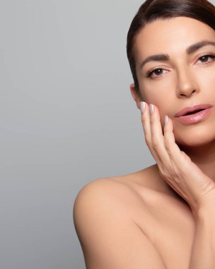 Yüzüne pürüzsüz bir cilt değdiren, doğal güzelliği ve cilt konseptini somutlaştıran orta yaşlı muhteşem bir kadın. Kopyalama alanı olan gri bir arkaplanda izole edilmiş.