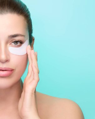 Cilt bakımı için göz altı bandı kullanan bir kadının portresi. Turkuaz arka plan ferahlatıcı bir his katıyor. Geniş kopya alanı ile izole edilmiş.