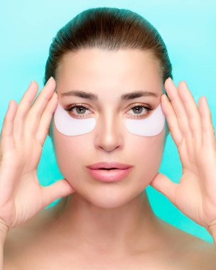 Cilt bakımı için göz altı bandı kullanan bir kadının güzelliği ve sağlığı için yakın plan çekimi. Kopyalama alanı olan mavi bir arkaplanda izole edilmiş.