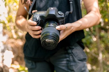 DSLR kameralı tanınmayan bir kadın iki elini de açık havada tutuyor. Kameraya odaklanmış durumda, fotoğrafçılık kavramları ve açık hava keşifleri vurgulanıyor. Orta bölüm yakın çekim
