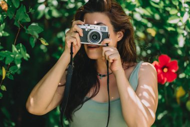 Eski tarz bir kamera kullanarak yemyeşil bir bahçede fotoğraf çeken bir kadın. Güneş ışığı yüzünde desenler oluşturur. Arka planda parlak kırmızı çiçek. Resim dikey biçimdedir.
