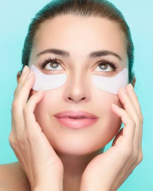 Cilt bakımı için göz altı bandı kullanan bir kadının yakın çekimi. Mavi bir arka plan karşısında rahatlamış ve yenilenmiş görünüyor. Bu da güzelliği ve sağlığını gösteriyor. Kopya alanı ile izole edilmiş arkaplan.