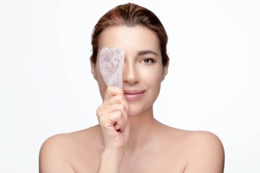 Huzurlu bir kadın, yüz masajı için gua sha aracı gösterir, kopya alanı olan beyaz bir arka planda cilt bakımını ve sağlığını teşvik eder.