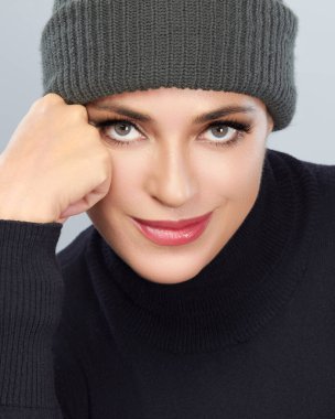 Örgü şapka ve kazak giyen, neşeli bir ifade sergileyen bir kadının yakın plan portresi. Yumuşak ışıklandırma ve nötr arkaplan resmi güçlendirir.