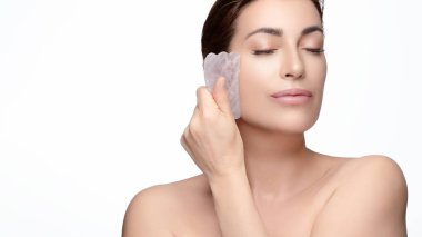 Huzurlu bir kadın, yaşlanma karşıtı cilt bakımı için yüzüne gua sha aleti ile nazikçe masaj yapıyor. Görüntü doğal güzelliği ve kendine bakımı somutlaştırıyor. Geniş kopya alanı olan beyaz bir arkaplanda izole edilmiş.