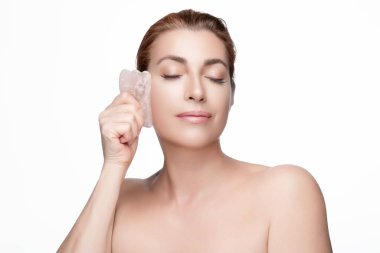 Huzurlu bir kadın yüz masajı için gua sha gül kuvarsı kullanır, güzellik ve cilt bakımı kavramlarını ön plana çıkarır. Kopyalama alanı olan hafif bir arkaplanda izole edilmiş.