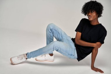 Yakışıklı Afrikalı Amerikalı adam gri arka planda siyah tişört ve mavi kot pantolon giyiyor.