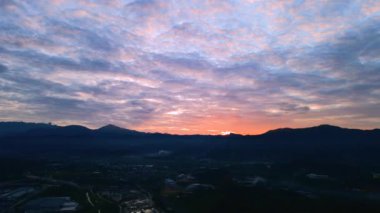 4K insansız hava aracı görüntüleri, turuncu, mor gökyüzü ve puslu tepelerle endüstriyel bir arazide gün doğumunu gösteriyor, sakin, sinematik bir manzara sunuyor..