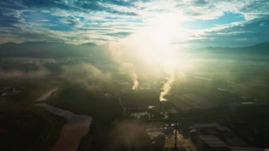 4K hava görüntüsü, fabrika binaları, bacalar, sisli manzaralar ve mavi gökyüzünün altındaki dağlar ile gündoğumunda bir sanayi arazisi..