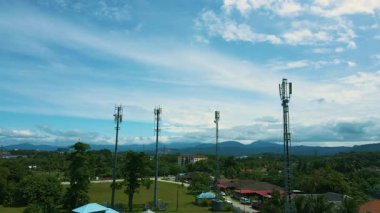 Yeşil ve uzak dağlarla çevrili bir banliyö bölgesinin üzerinde yükselen 5G antenlerinin havadan 4K görüntüsü. Haberleşme ve teknoloji projeleri için ideal.