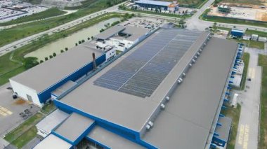 Çatıda güneş panelleri olan büyük bir endüstriyel binanın havadan 4K görüntüsü. Etrafı yollar, bir gölet ve yeşilliklerle çevrili. Temiz enerji ve sürdürülebilirliği teşvik eder.