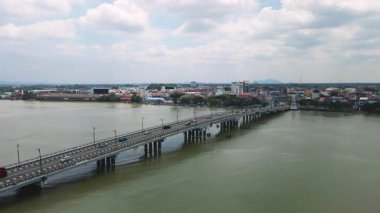 Hareketli araçlarla sakin bir nehri geçen uzun bir köprünün havadan görüntüsü. Köprü, bulutlu bir gökyüzünün altındaki binalar, yollar ve ağaçlarla gelişmiş bir kasaba alanını birbirine bağlıyor..
