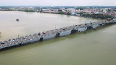 Hareketli araçlarla sakin bir nehri geçen uzun bir köprünün havadan görüntüsü. Köprü, bulutlu bir gökyüzünün altındaki binalar, yollar ve ağaçlarla gelişmiş bir kasaba alanını birbirine bağlıyor..
