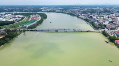 Malezya 'da geniş bir nehrin üzerindeki uzun bir köprünün havadan görünüşü. Tekneler ve araçlar geçiyor. Arka planda şehir manzarası.