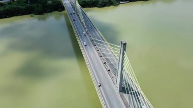 Orman ve binalarla çevrili geniş bir nehri geçen kablo destekli uzun bir köprünün havadan görüntüsü. Modern altyapı.