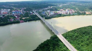 Orman ve binalarla çevrili geniş bir nehri geçen kablo destekli uzun bir köprünün havadan görüntüsü. Modern altyapı.