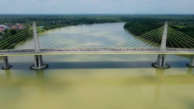 Orman ve binalarla çevrili geniş bir nehri geçen kablo destekli uzun bir köprünün havadan görüntüsü. Modern altyapı.