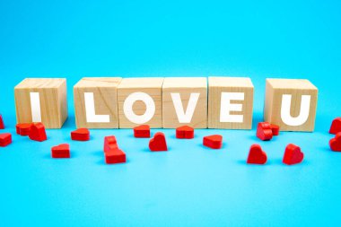 Wooden cubes arranged to spell I LOVE U with small red heart shapes scattered on a blue background. Romantic love message concept for Valentines Day, relationships, or celebration of affection and care.