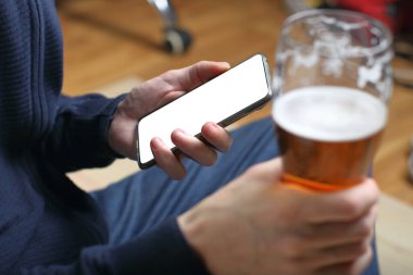 a person is holding a smartphone and a mug of beer in his hands while sitting cross-legged
